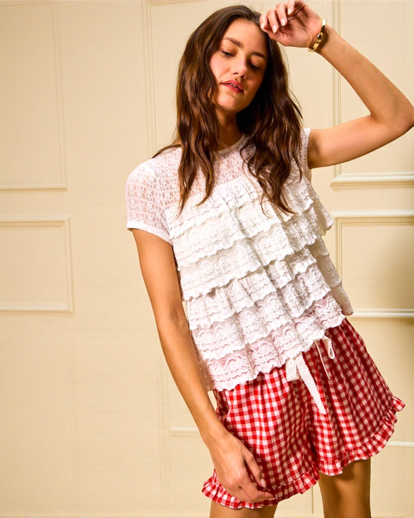 Woman wearing a white lace top and red checkered shorts against a beige wall.