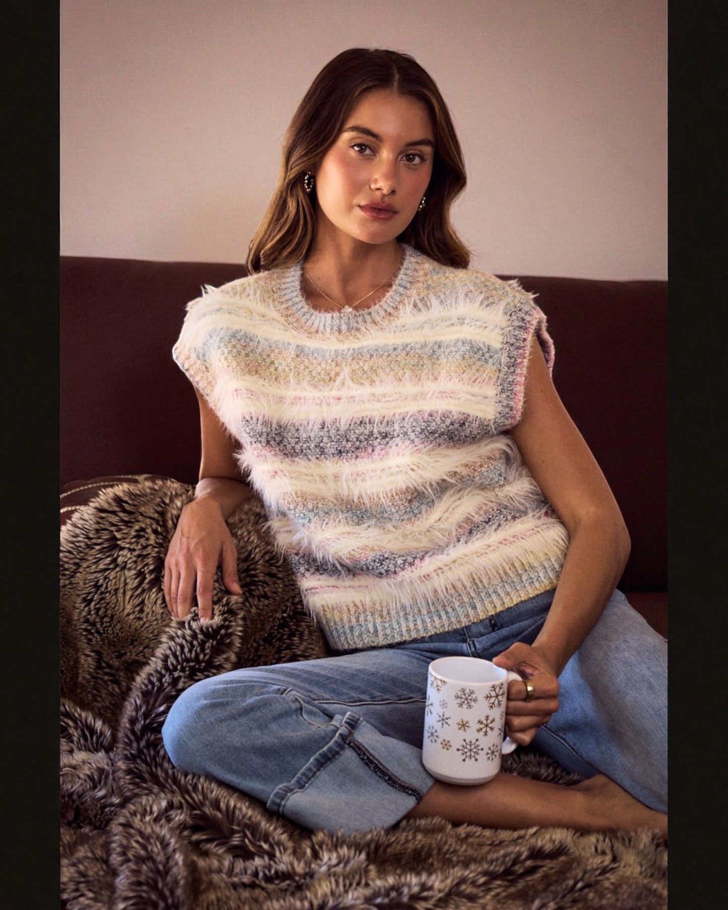 Woman wearing a striped sleeveless sweater sitting on a couch with a mug.