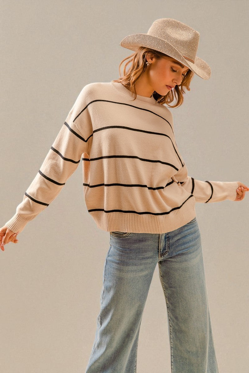 Woman wearing a beige sweater with black stripes, light blue jeans, and a beige hat against a plain background