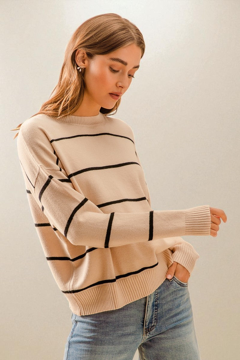 Woman wearing a beige sweater with black stripes and light blue jeans against a plain background