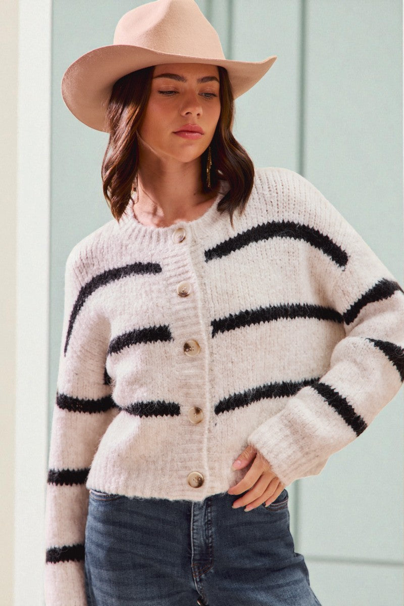 Woman wearing a cream cardigan with black stripes, blue jeans, and a beige hat against a light green background.
