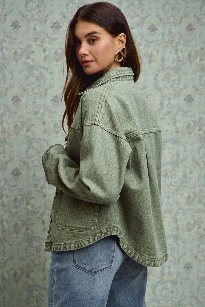 Woman wearing a green denim jacket and blue jeans against a floral-patterned wall.