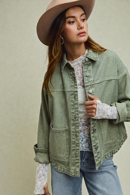 Woman wearing a green denim jacket, white lace shirt, and blue jeans with a beige hat against a neutral background.