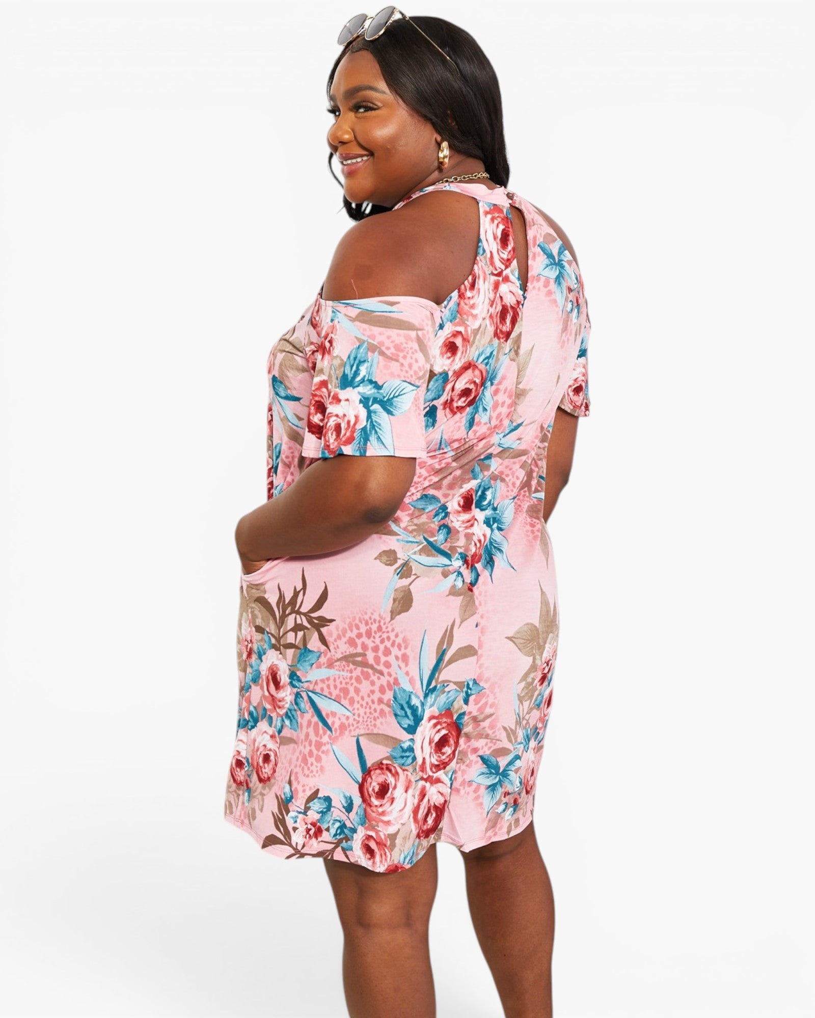 A woman modeling a pink floral cold-shoulder dress with blue and red flowers, shown from a three-quarter back angle against a light gray studio background.