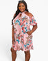 A woman modeling a pink cold-shoulder mini dress with a floral print featuring red, blue, and brown flowers on a light pink background, set against a plain light gray studio backdrop.