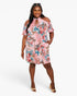 A woman modeling a pink cold-shoulder mini dress with a blue, red, and brown floral print, styled with beige high-heeled sandals and sunglasses on a light gray background.