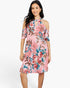 A woman modeling a pink cold-shoulder midi dress with a vibrant floral print in shades of red, blue, and brown, set against a plain white studio background.
