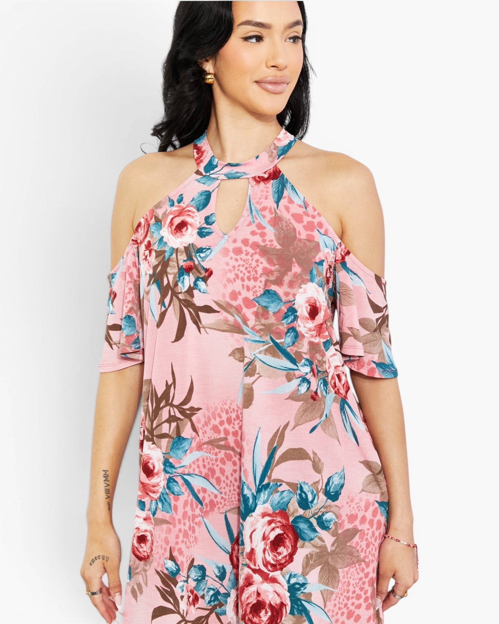 A woman wearing a pink cold-shoulder floral dress with blue, red, and brown botanical print details, shown against a plain light gray studio background.