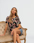 Woman sitting on a vintage couch wearing a patterned top and denim shorts against a plain background