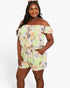 A woman modeling a light green off-the-shoulder floral romper with a tie waist and side pockets, photographed against a plain light gray studio background.