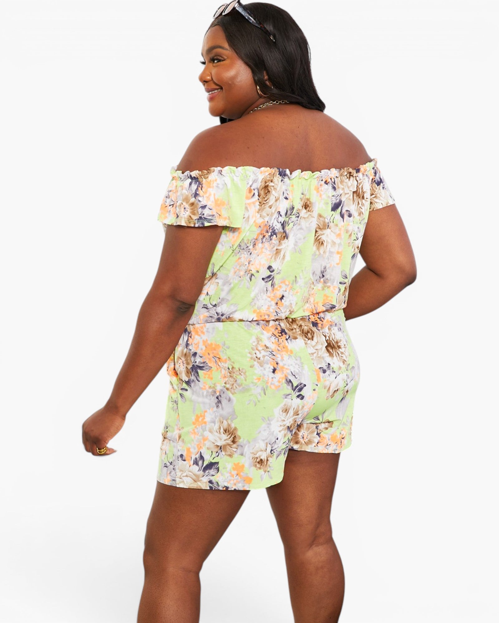 A woman modeling an off-the-shoulder floral romper from the back, featuring a light green base with orange, yellow, and white floral patterns, against a plain white background.