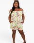 A woman modeling a light green off-the-shoulder floral romper with orange, yellow, and blue flowers, set against a plain white studio background.