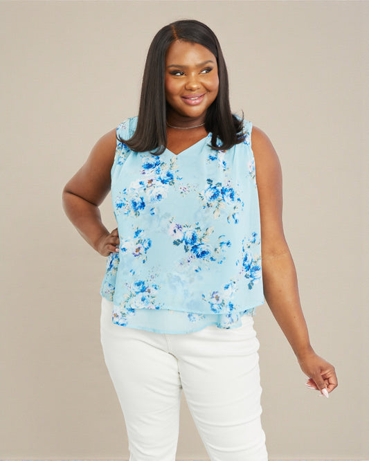 Woman wearing a light blue floral sleeveless top and white pants on a beige background