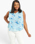 A woman modeling a sleeveless light blue blouse with a blue and white floral pattern, paired with white pants, against a plain white studio background.