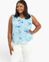 A woman modeling a sleeveless light blue V-neck blouse with a blue and white floral pattern, paired with white pants, against a plain white background.
