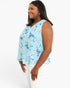 A woman wearing a light blue sleeveless blouse with a blue, white, and pink floral pattern, paired with white pants, photographed against a solid white studio background.