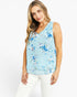 A woman modeling a light blue sleeveless top with a blue and white floral pattern, paired with white pants, against a plain white background.