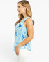 A woman shown in side profile wearing a light blue sleeveless top with a blue and white floral pattern, paired with white pants, against a plain white studio background.