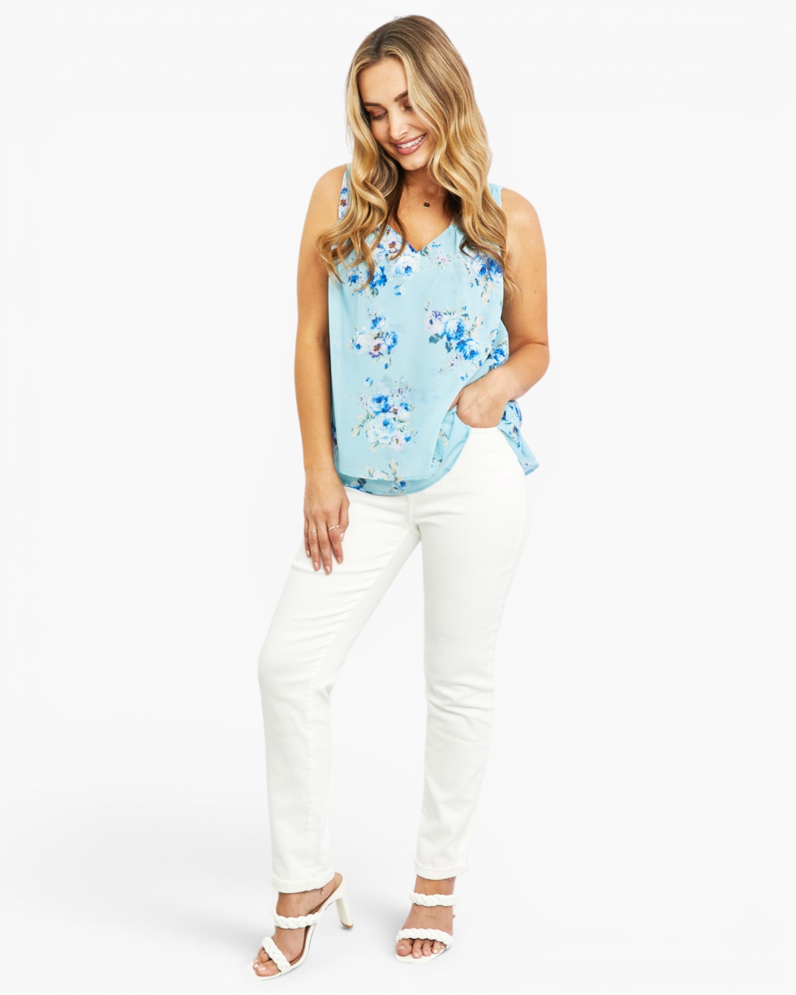 A woman modeling a light blue sleeveless top with a blue and white floral print, paired with white pants and white heeled sandals, against a plain white studio background.