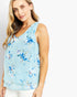 A woman wearing a light blue sleeveless top with a blue, purple, and white floral print, paired with white pants, photographed against a plain white background.