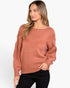 A woman wearing a dusty rose knit sweater with textured lace-like yoke and sleeve details, paired with black pants, against a plain white studio background.