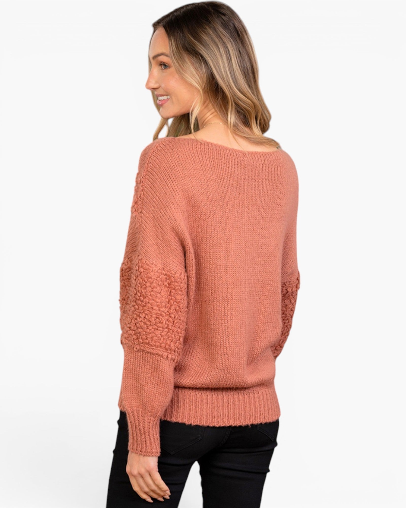 A woman wearing a dusty rose knit sweater with textured, chunky knit elbow patches, shown from a three-quarters back view against a plain white background.