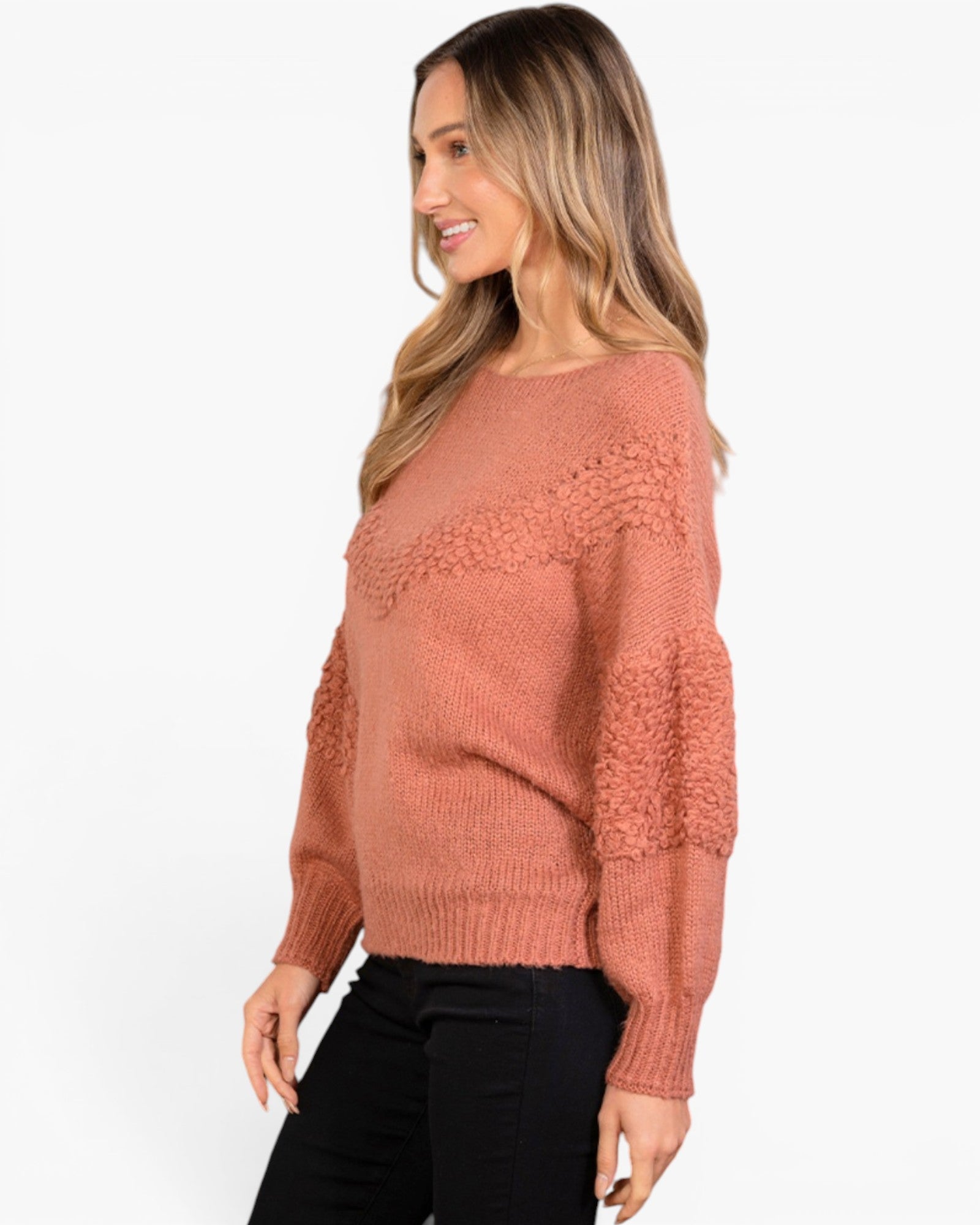A woman wearing a dusty rose knit sweater with textured cable details on the sleeves, paired with black pants, shown from a side profile against a plain white background.