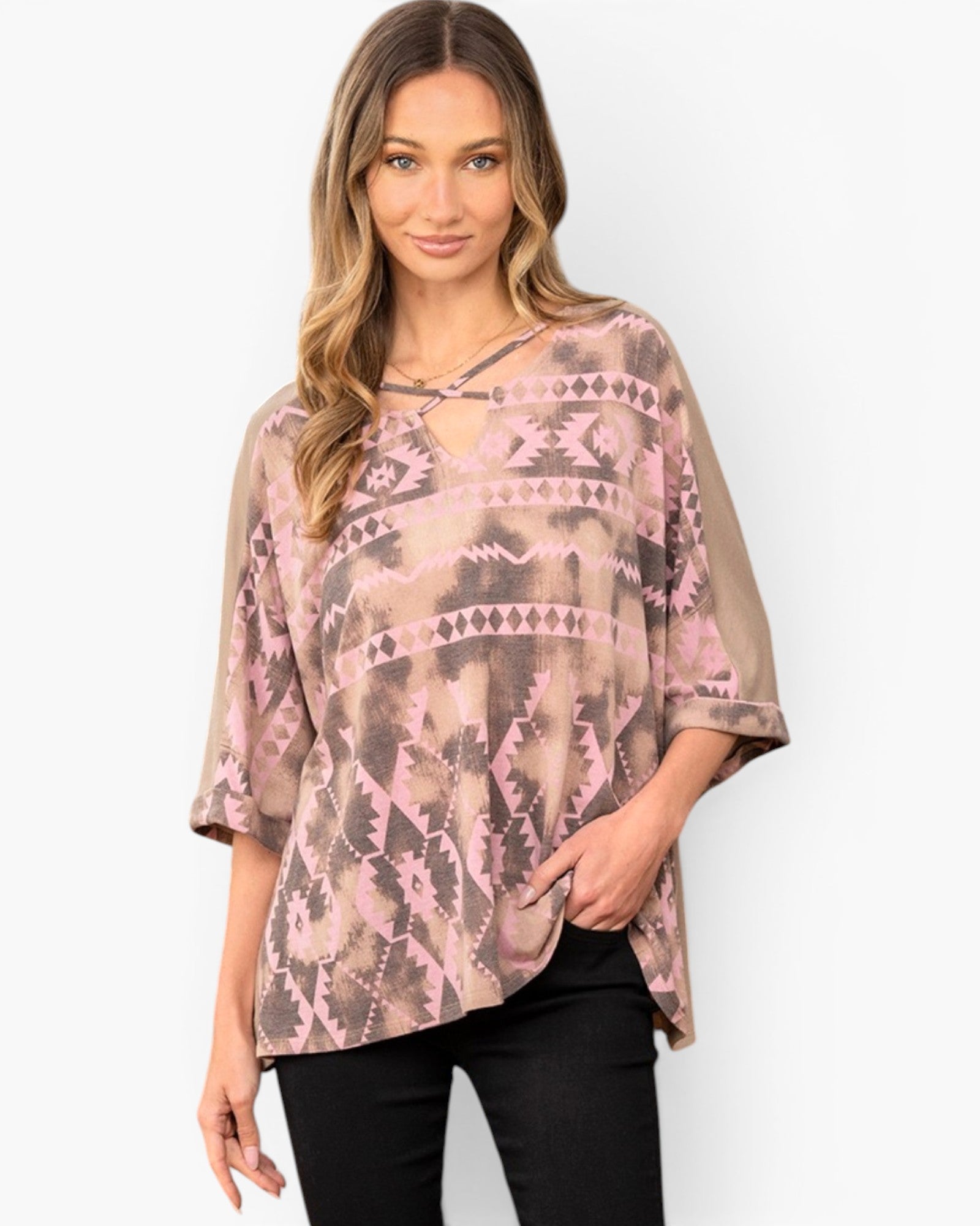 A woman modeling a loose, short-sleeve top with a pink and taupe geometric tribal pattern, paired with black pants, against a light gray studio background.