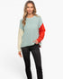 A woman modeling a color-block knit sweater with a sage green body, cream sleeves, and a bright red sleeve, paired with black pants against a light gray studio background.
