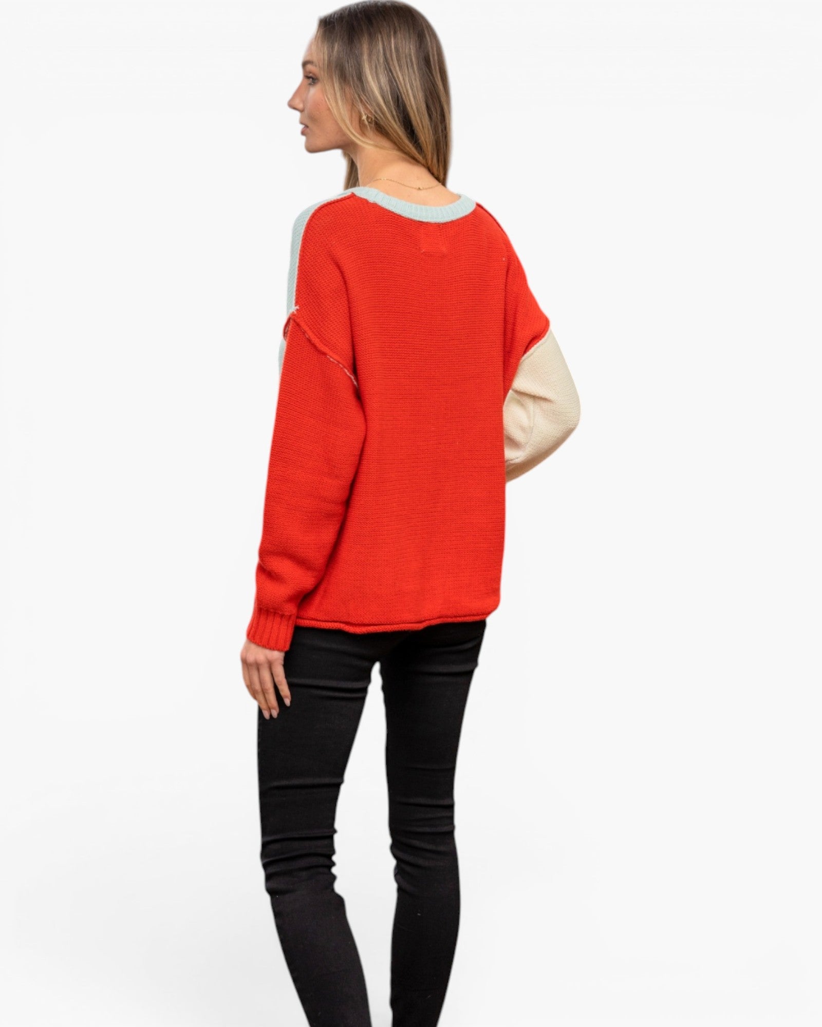 A woman shown from the back wearing a bright red knit sweater with contrasting light beige sleeves and a light gray crew neck, paired with black pants, against a plain white background.