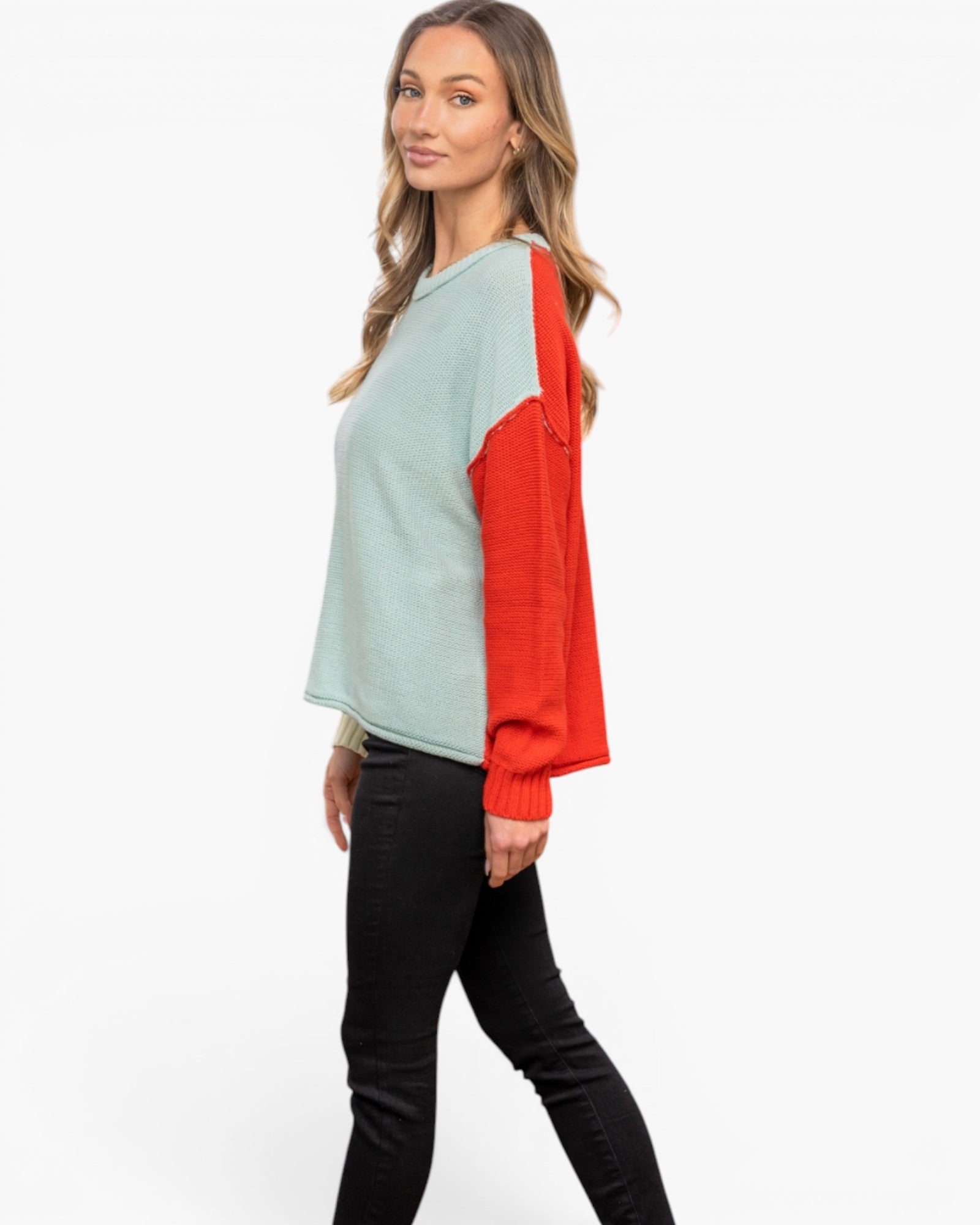 A woman modeling a colorblock knit sweater with a light sage green body and bright red sleeves, paired with black pants, against a plain white studio background.