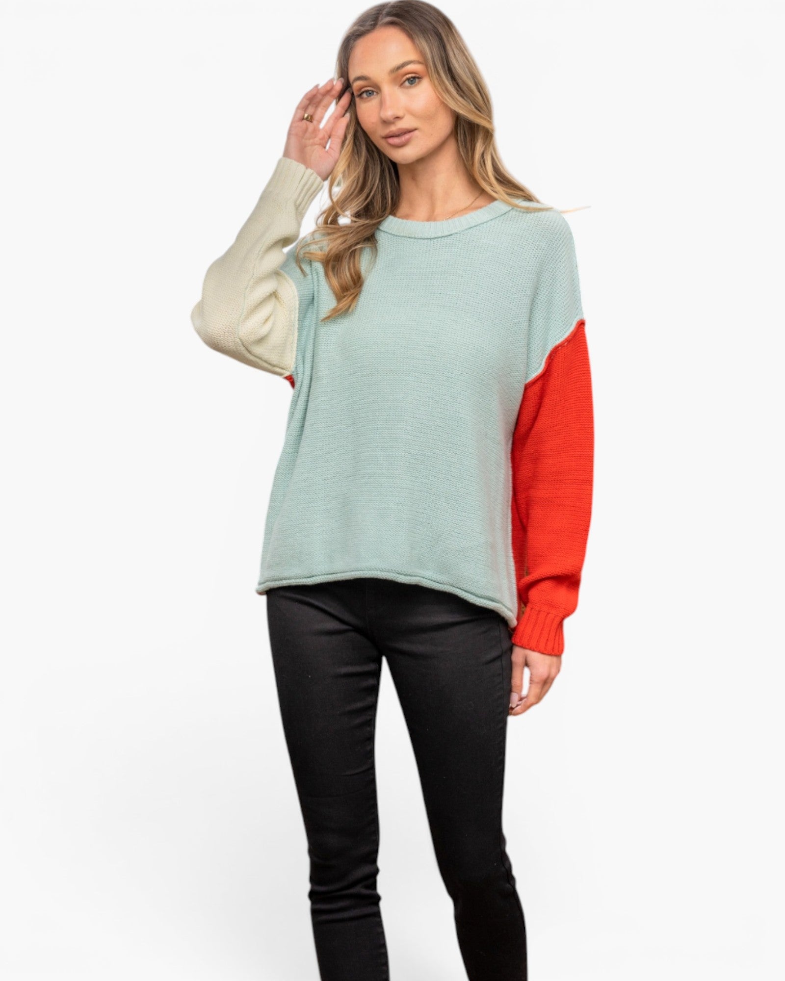 A woman modeling a color-block knit sweater with a light sage green body, cream-colored sleeves, and bright red color-blocked sleeves, paired with black pants against a plain white background.