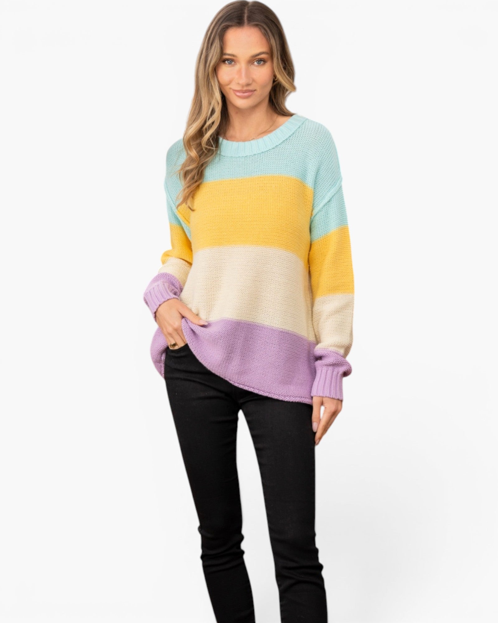 A woman modeling a fuzzy knit crewneck sweater with horizontal color-blocked stripes in light blue, yellow, cream, and lavender, paired with black pants against a plain white background.