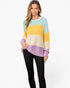 A woman modeling a fuzzy knit crewneck sweater with horizontal color-blocked stripes in light blue, yellow, cream, and lavender, paired with black pants against a plain white background.