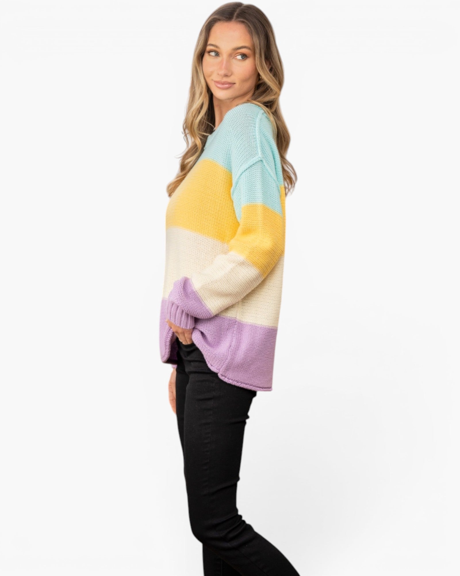 A woman wearing a colorful color-block knit sweater with horizontal stripes of light blue, yellow, cream, and lavender, paired with black pants, photographed against a plain white studio background.