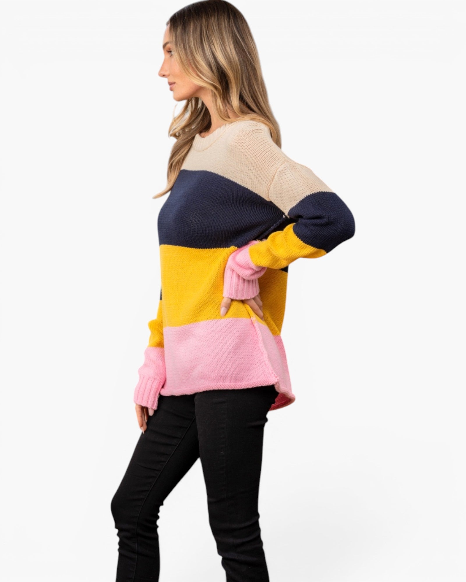 A woman wearing a color-block knit sweater with beige, navy, yellow, and pink sections, paired with black pants, shown against a plain white studio background.