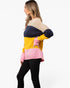 A woman wearing a color-block knit sweater with beige, navy, yellow, and pink sections, paired with black pants, shown against a plain white studio background.