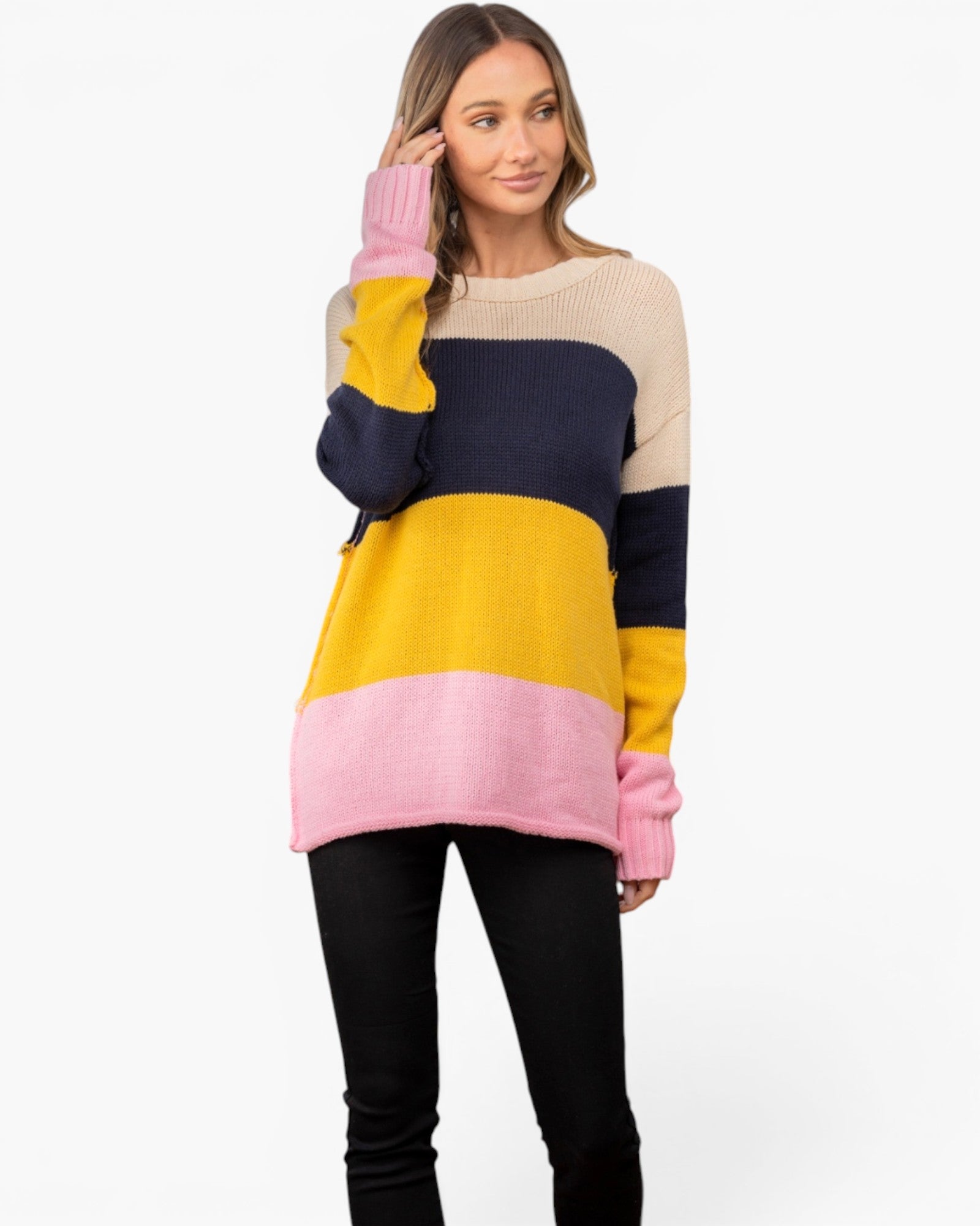 A woman modeling a color-blocked knit sweater with horizontal stripes of pink, yellow, navy, and beige, paired with black pants against a plain white background.