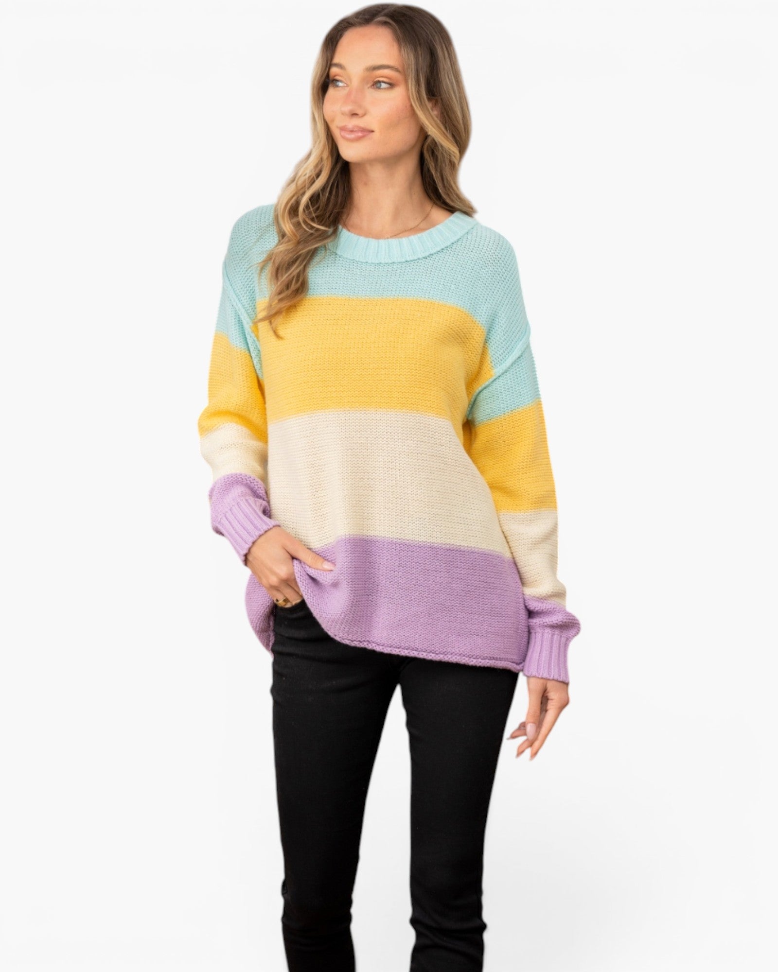 A woman modeling a color-blocked knit sweater with horizontal stripes of light blue, yellow, cream, and lavender, paired with black pants, against a plain white studio background.
