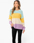 A woman modeling a color-blocked knit sweater with horizontal stripes of light blue, yellow, cream, and lavender, paired with black pants, against a plain white studio background.
