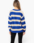 A woman viewed from the back wearing a blue and cream striped knit sweater and black pants, against a plain light gray studio background.
