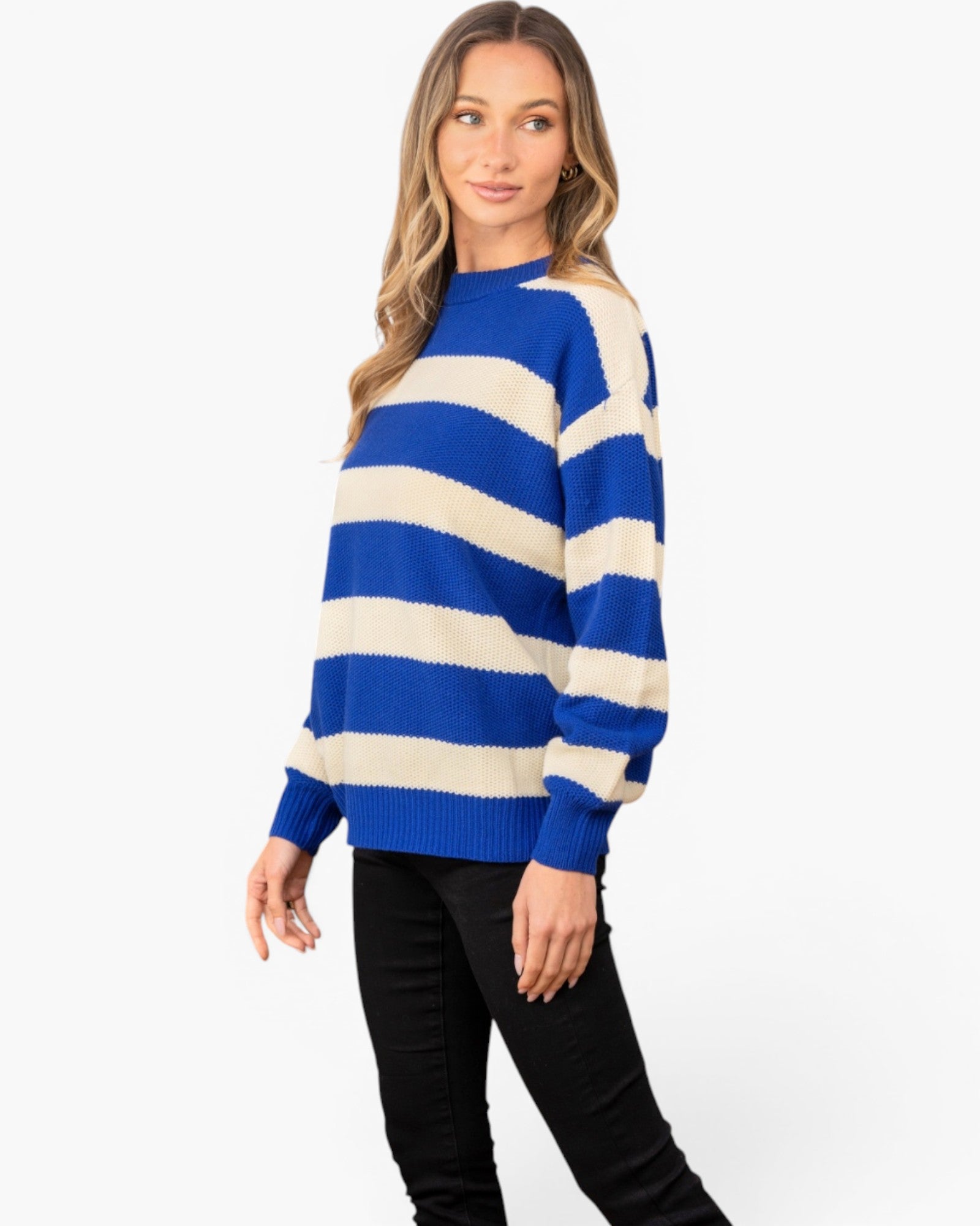 A woman modeling a blue and cream striped knit sweater with black pants, standing against a plain white studio background.