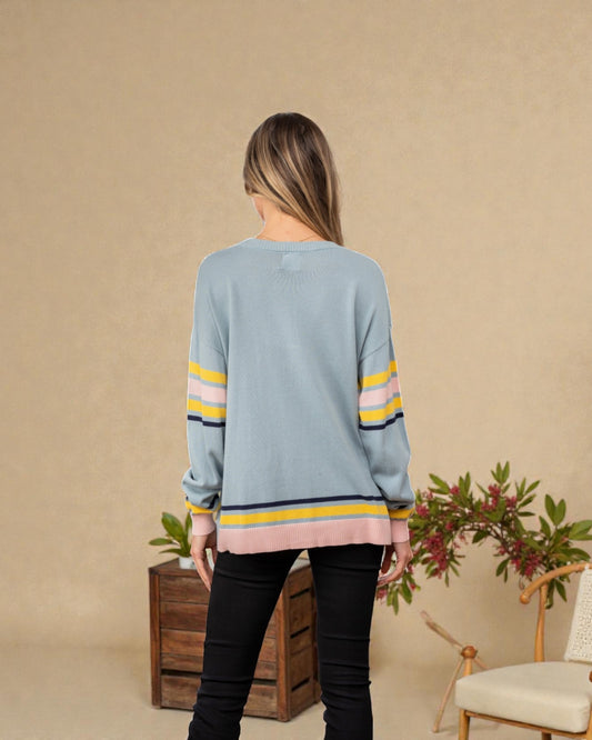 Woman wearing a light blue sweater with colorful stripes, standing against a beige wall with decorative elements.