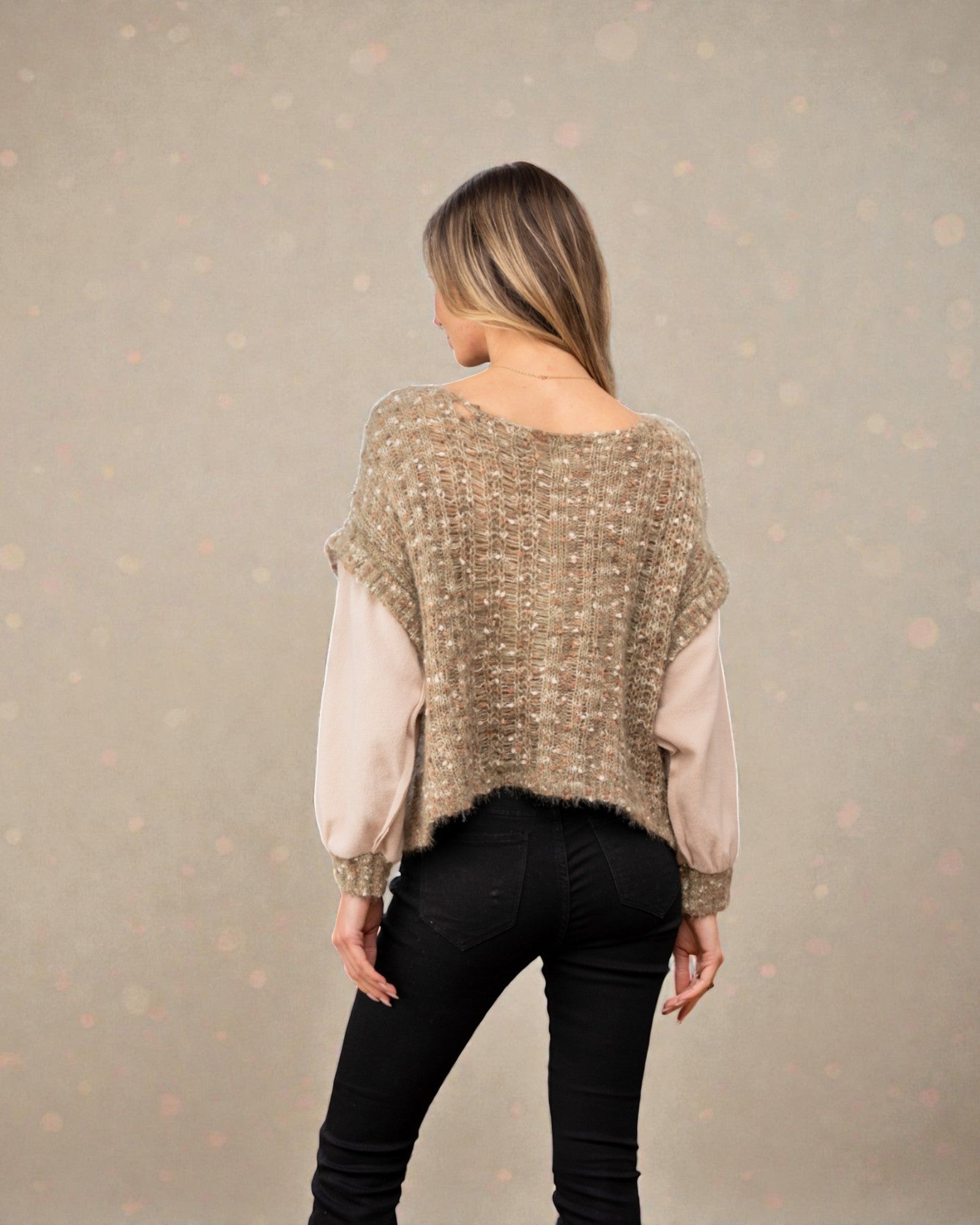 Person wearing a beige knitted poncho over a light pink shirt and black pants on a neutral background
