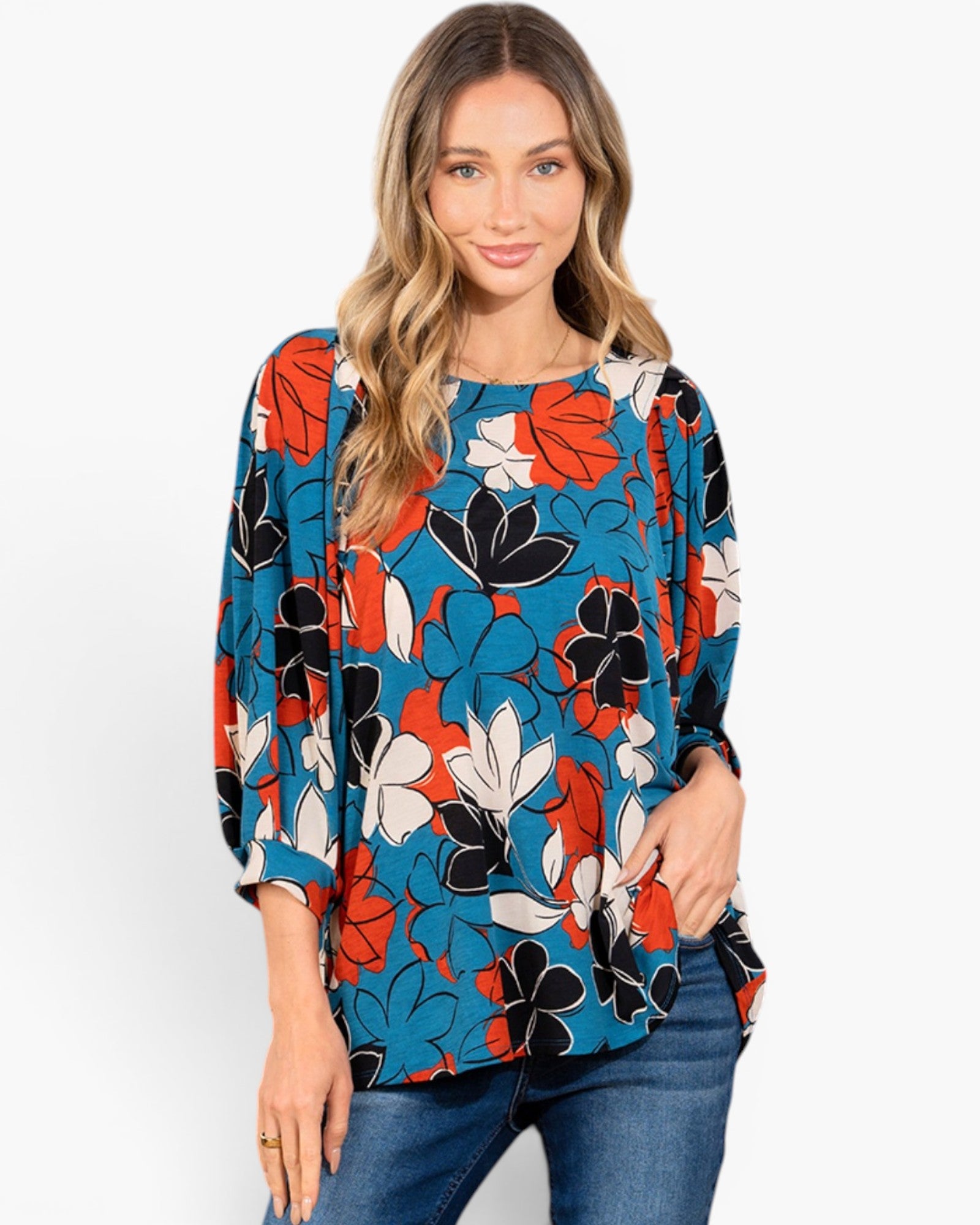 A woman wearing a blue long-sleeve blouse with a bold orange, black, and white floral pattern, paired with blue jeans, against a plain white background.