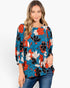 A woman wearing a blue long-sleeve blouse with a bold orange, black, and white floral pattern, paired with blue jeans, against a plain white background.
