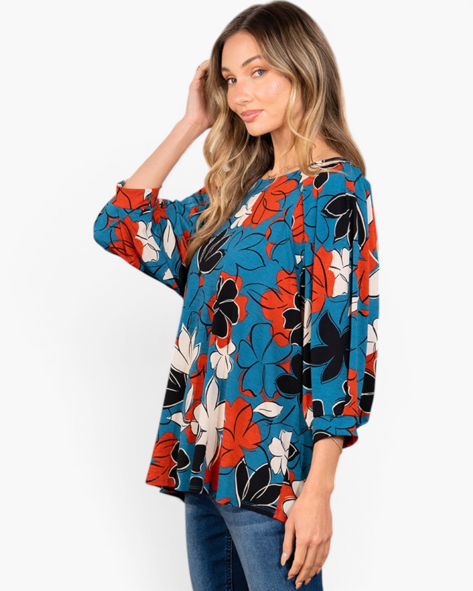A woman modeling a blue floral long-sleeve top with red, black, and white flowers, paired with blue jeans, against a plain white background.