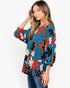 A woman modeling a blue floral long-sleeve top with red, black, and white flowers, paired with blue jeans, against a plain white background.