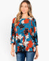 A woman wearing a vibrant floral long-sleeve top with blue, orange, black, and white flowers, paired with blue jeans, against a plain white background.