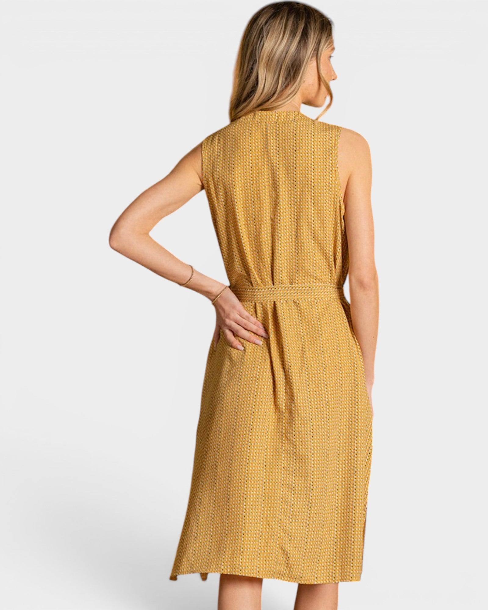 A woman shown from the back wearing a sleeveless midi dress with a textured, woven mustard yellow and white pattern. The dress features a high neckline and a matching fabric tie belt at the waist, set against a plain light gray studio background.